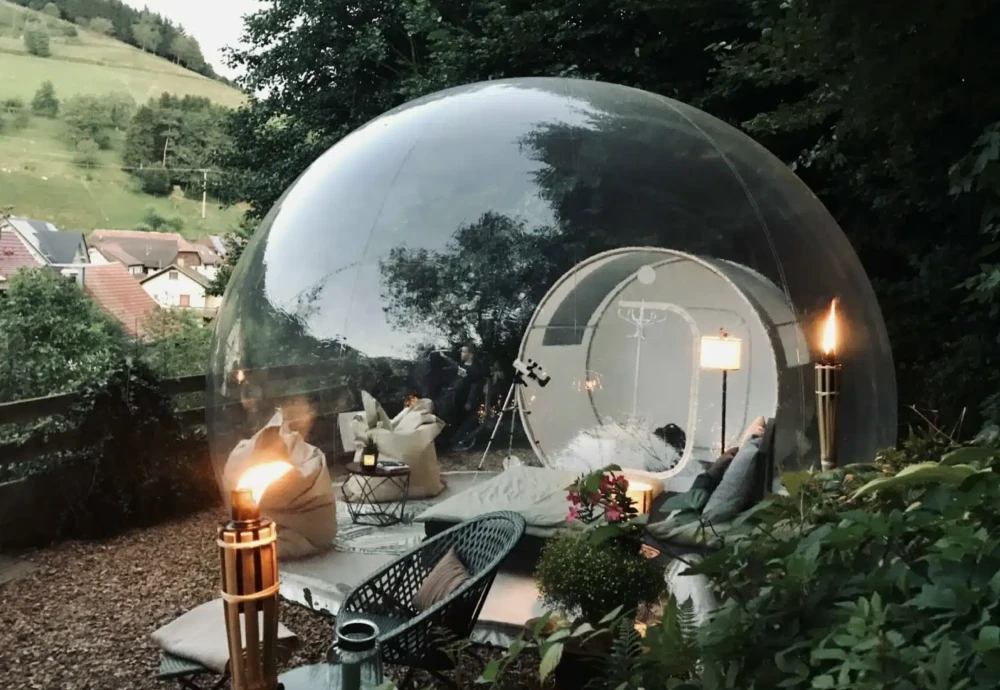 Transparent inflatable bubble tent set on a wooden deck overlooking a valley, furnished with cozy outdoor seating and soft lighting for daytime glamping

