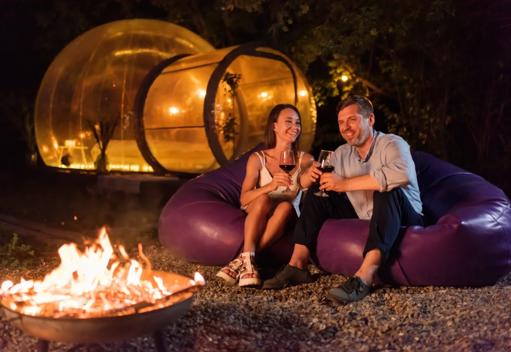 Couple enjoying wine by a campfire in front of an illuminated inflatable bubble tent during a glamping night outdoors