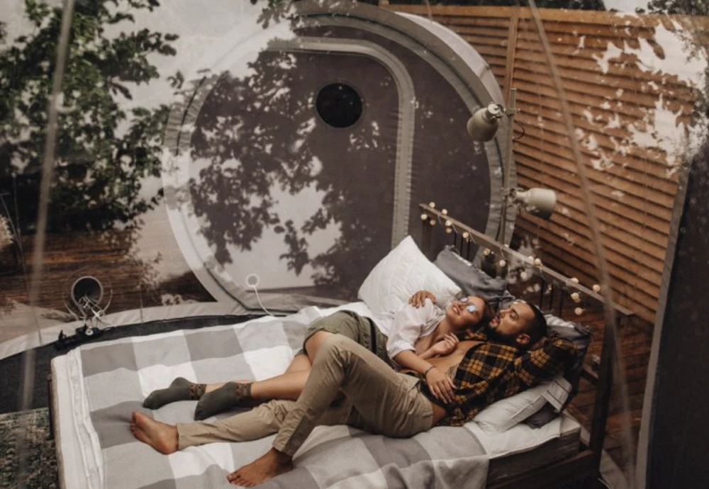 Couple lying on a bed inside a clear bubble tent during the day, surrounded by natural light and trees for a romantic and intimate glamping moment

